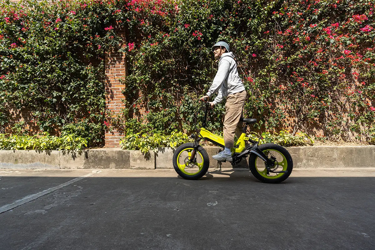 MIHOGO NX lime yellow foldable fat tyre electric bike, rider wearing helmet cycling beside floral brick wall, compact folding e-bike for city commuting and outdoor leisure rides